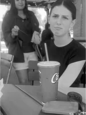 A black and white photo features a woman seated at a restaurant table, with a soda cup in front of her. She gazes directly at the camera with a look of slight displeasure, her expression suggesting betrayal or annoyance. Behind her, standing near the restaurant's entrance, is another woman who appears to be walking away. The overall composition is simple, with the subjects in focus against a blurred background.