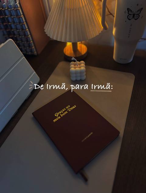 A softly lit, top-down shot of a bedside table or desk. A burgundy-colored journal lies open, with gold text. Adjacent to it is a light-colored, rectangular mousepad. A cylindrical water bottle with a butterfly design sits further back, near the edge, with a small, bubbly candle, and a petite lamp with a white lampshade. Additional items on the desk and table provide a sense of a cozy, workspace.