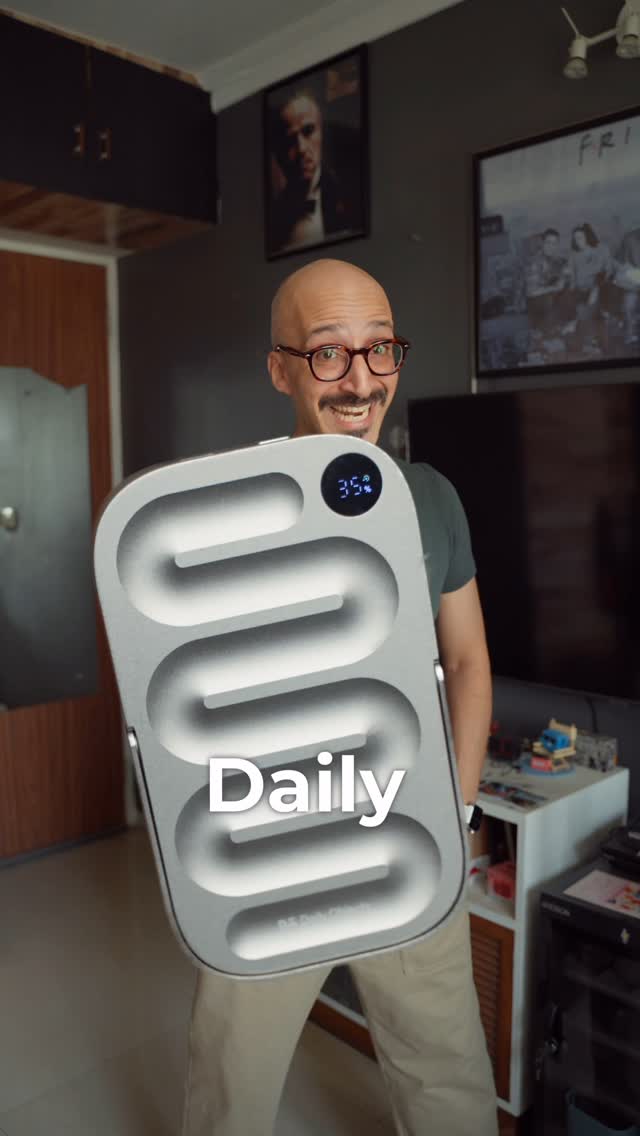 A man with glasses and a beard, holding a silver power bank labeled DailyObjects LOOP in front of him. He is smiling. The background includes a framed picture resembling a painting, a TV, and other home decor items. A digital display on the power bank shows the percentage of charge.