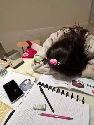 The image shows a person with long dark hair lying face-down on a cluttered desk, presumably studying. A pink illustration of a brain is added near the head. The desk is covered with papers, notebooks, a smartphone, a pen, an eraser, and a stapler. The composition suggests a study environment, and the person looks exhausted or overwhelmed. The background features a wall and part of a wooden chair.