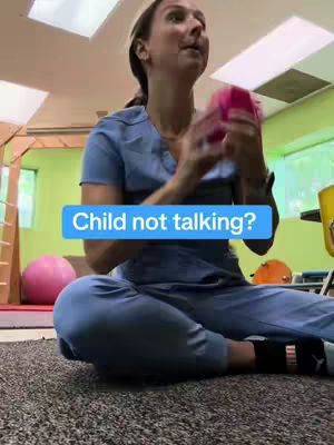 A woman in blue scrubs sits cross-legged on a carpeted floor, holding a pink object. The background shows a playroom with colorful accents.