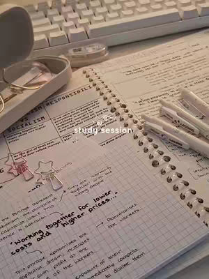 An overhead shot of a desk with a notebook open, pens, glasses case and a keyboard in the background. The open notebook contains handwritten notes and grid lines.