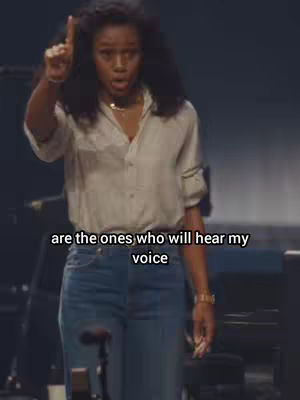 The image shows a woman with dark curly hair, wearing a light button-down shirt and blue jeans, standing with her right arm raised and index finger pointed upward. She appears to be speaking with an earnest expression, set against a dark background. Text overlayed at the bottom conveys a spiritual message.
