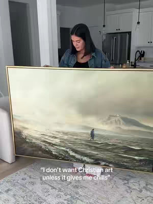 A woman in a denim jacket is holding up a large framed painting of a person walking on water in a stormy sea, the painting is positioned in front of the woman obscuring most of her body and face. In the background we can see a kitchen, and the room is well-lit. At the bottom of the image there is a grey rug with an abstract pattern.