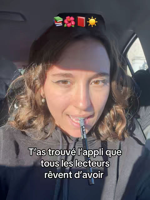 A young woman with light skin and curly brown hair is seated inside a car. The image is a medium shot, focusing on her face. She's slightly smiling, with a pen or similar object held between her teeth. She's wearing a dark hoodie. Above her head, a series of emojis are visible: a stack of books, a hibiscus flower, a red book, and a sun. The car interior, including a seat and a partially visible window, form the backdrop. The lighting appears to be natural.