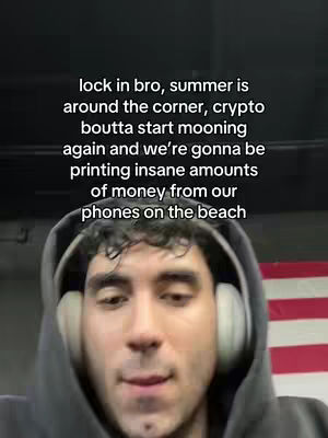A man wearing a gray hoodie and white headphones is positioned in the foreground. The background features a partial view of a red and white American flag. Text overlayed at the top reads: 'lock in bro, summer is around the corner, crypto boutta start mooning again and we're gonna be printing insane amounts of money from our phones on the beach'.
