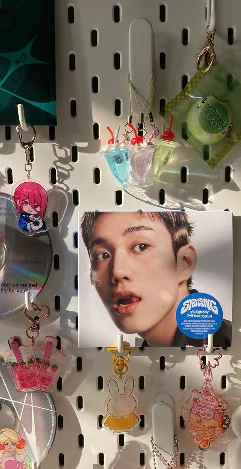 The image showcases a white pegboard adorned with various keychains, a framed picture of a young man, and small decor items. The keychains include colorful acrylic charms like a bunny, drink cups, and anime characters. The central image features a close-up of a man with his tongue out. The items create a layered, textured image with a bright, casual aesthetic.