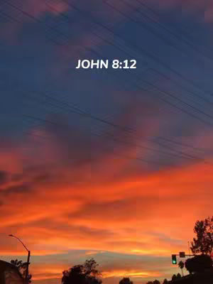 A scenic image of a sunset. The sky transitions from deep blue at the top to vibrant orange and red hues towards the horizon. Thin power lines are visible diagonally across the upper part of the sky. In the foreground, there are silhouetted trees and a street light, with the faint outlines of buildings. The text "JOHN 8:12" is overlaid in white.