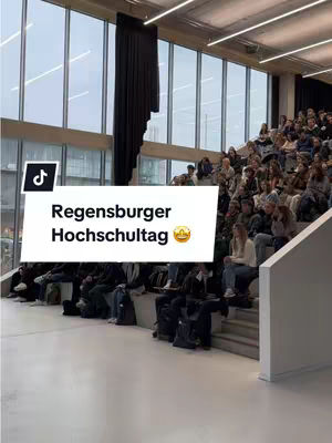 A wide shot of a large auditorium filled with students seated on tiered seating. Large windows span the back wall, letting in natural light and offering an outside view. A black curtain hangs on the left side of the windows. In the foreground, a white text box with the words, 'Regensburger Hochschultag' is overlaid with the TikTok logo on the left, with a crown emoji at the end.