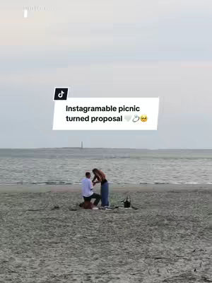 A couple on the beach, with the man kneeling in front of the woman proposing, a picnic setup is near them. Overcast sky over the ocean in the background.