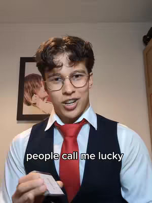 A young man in a dress shirt, vest, and tie smiles while holding a small card. The phrase "people call me lucky" is displayed at the bottom of the frame. A framed picture hangs in the background.