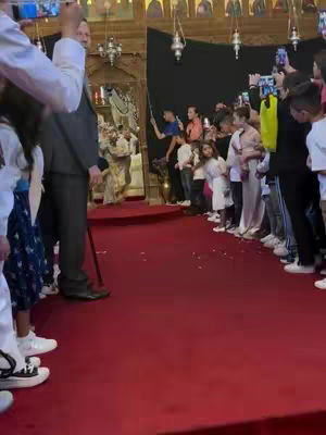 The scene is of a Greek Orthodox Church interior. A red carpet leads to a raised altar area. A man in a gray suit and a black clergy member are in focus, along with many people. Some are recording the scene on their phones.
