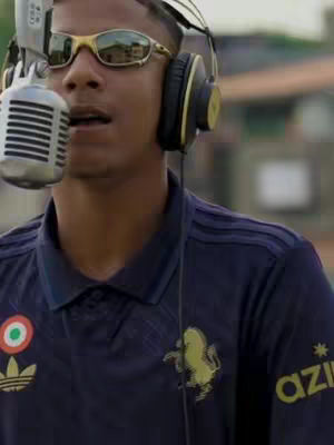 The image showcases a young man in a close-up, singing into a microphone. He wears a blue Adidas polo shirt with a gold logo, gold-rimmed sunglasses, and headphones. The background is slightly blurred but shows a city backdrop.