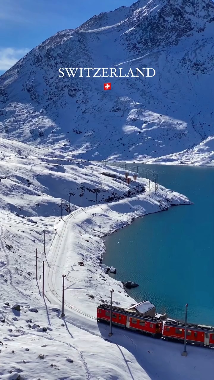 An aerial shot of a snow-covered landscape featuring a turquoise lake, a winding road, and a train. The mountains are covered with snow, and the sky is clear