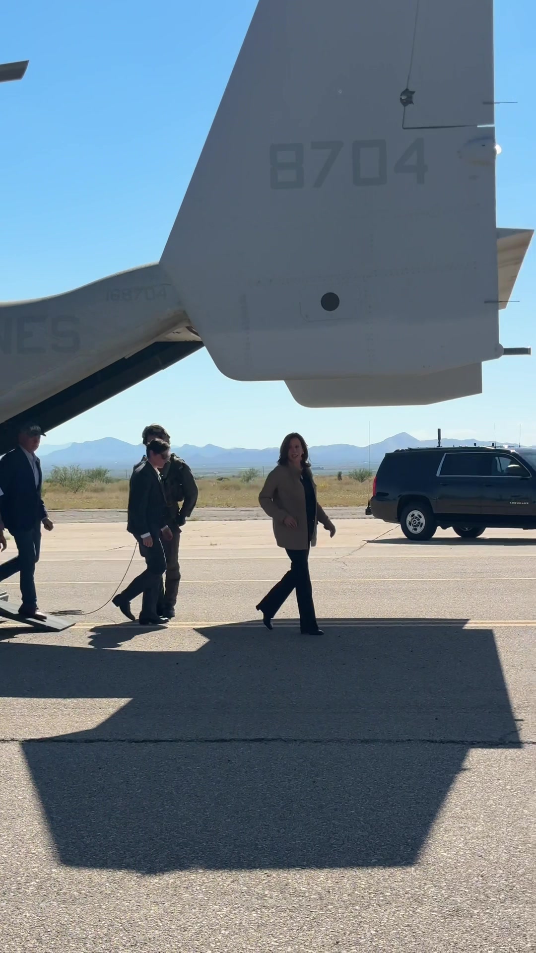 The cover image shows a group of people walking on a paved surface, likely an airport tarmac. In the foreground, a woman in a tan jacket and black pants is walking, with the tail of a military aircraft in the background. Several other people, including men wearing suits, are walking alongside her, and a black SUV is parked nearby.