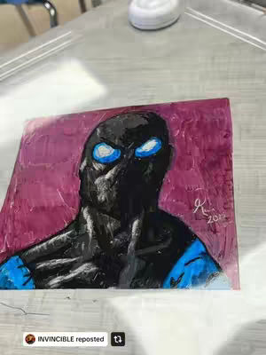 A close-up drawing of a character from the show Invincible. The character has a black masked head with blue eyes painted on a pink background. The work is displayed on a surface and signed at the bottom right.