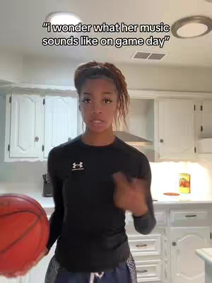 A young Black woman stands in a kitchen, holding a basketball. The background features white cabinets, a stove hood, and a round ceiling light. The image is well-lit, with the woman as the central focus, posed indoors.