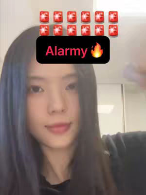 A young woman with long black hair is the primary focus, smiling slightly towards the viewer. Above her head are small, red alarm clock icons arranged in two rows. Overlaying her forehead is a black rectangle containing the word "Alarmy" in pink, with a flame emoji next to it. The background is simple and slightly blurred, suggesting an indoor setting.