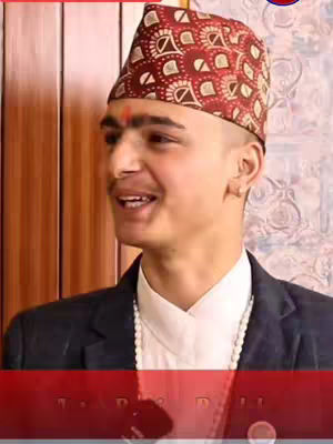 A man with short hair, wearing a patterned cap, suit jacket, and a white shirt, smiles while looking slightly off-camera. There is a visible red design on the lower portion of the image. The setting appears to be indoors, with a wooden panel background.