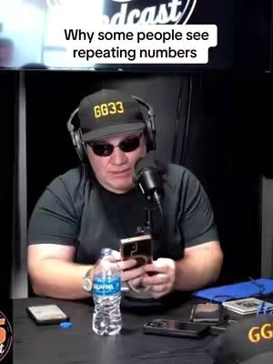 A man wearing sunglasses, a hat, and a headset sits at a desk with a microphone. He is holding a phone and there is a bottle of water and other items on the desk. The text overlay states, Why some people see repeating numbers.