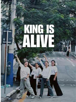 The image features four people on a city street, with a dramatic sky and trees in the background. The words 'KING IS ALIVE' are prominently displayed in large, white block letters in the center. The people are positioned below the text, raising their arms, with expressions that convey energy.