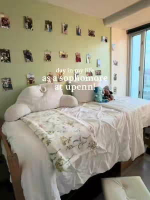 The cover image shows a bedroom scene. The bed is made with white bedding, and a patterned throw is draped across the foot of the bed. A large, cloud-shaped pillow sits atop the bed. The wall behind the bed is painted a light green and decorated with numerous framed photos. Text overlay on the image reads 'day in my life as a sophomore at upenn!'.