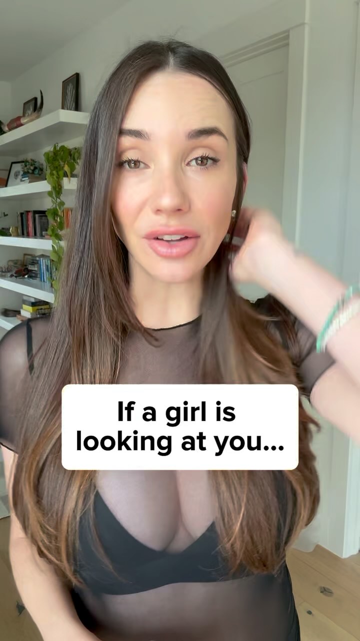 A woman with long brown hair is the main subject. She's looking slightly upwards with a neutral expression. A white rectangle with black text is positioned at the bottom middle of the frame. She is wearing a black top, and the background shows a white wall with some shelves with books.