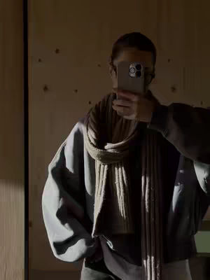 A person is taking a picture with their phone in a mirror. They are wearing a scarf and a jacket. The background has wooden textures.
