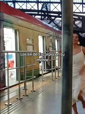 A woman in a white dress stands on a train platform, smiling and looking at the camera. Behind her, a train is visible, along with the platform's metal railings and overhead supports. The background is blurred, suggesting movement or depth.