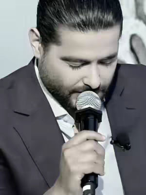 The image is a medium shot of a man with a beard and dark hair, holding a microphone. He is dressed in a suit with a white shirt. The man is looking down, seemingly in concentration or prayer. The background is out of focus, suggesting a close-up. The overall lighting is muted, with a focus on the man's face. The microphone is the central element.