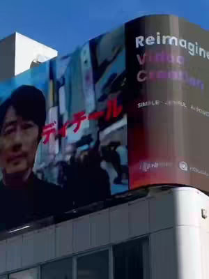 An urban outdoor scene featuring a large billboard with Japanese text and an image of a man. The billboard is positioned atop a building with a clear blue sky in the background.