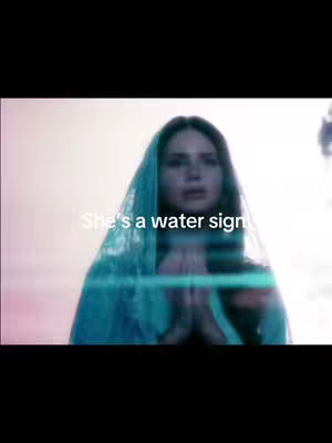 A woman in a blue veil-like scarf is the focus, with the text overlayed reading "She's a water sign". The background is blurred, with a soft, ethereal glow adding to the dreamy quality of the composition.