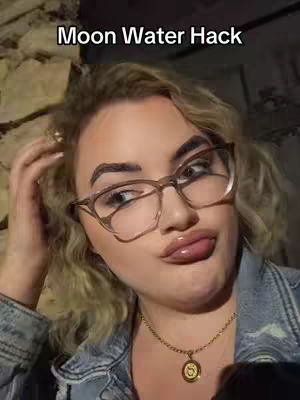 A woman with blonde curly hair and glasses purses her red lips, touching her hair, with the text 'Moon Water Hack' overlaid on top. She wears a necklace with a pendant and a denim jacket.