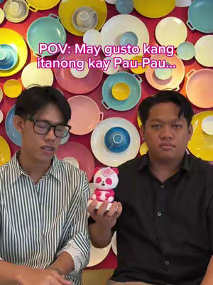 Two men pose in front of a wall decorated with plates, holding a panda figurine. The man on the left wears glasses and a striped shirt, and the man on the right wears a black shirt. They both have neutral expressions. The background consists of varied colored plates.