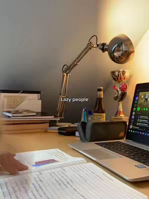 A desk setup with a laptop, open notebook, and lamp. The scene is lit with warm light. A bottle and trophy are visible behind the laptop.