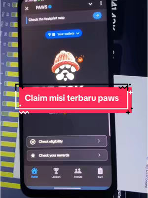 The image shows a smartphone screen displaying an app interface. The screen is black with a red and white banner overlayed. The banner reads "Claim misi terbaru paws". The background consists of a blurred view of an interior with some grid structures and other tech devices. The app interface has the PAWS logo on the top with a paw print design and other icons along the bottom.