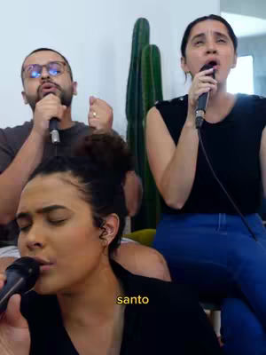 The image shows three people, likely a singing group, in a well-lit indoor setting. In the foreground, a woman with dark curly hair is singing into a microphone, and a smaller text overlay of the word "santo" appears near her. Behind her, sitting on couches, are two others singing into microphones, an older man wearing glasses and another woman.