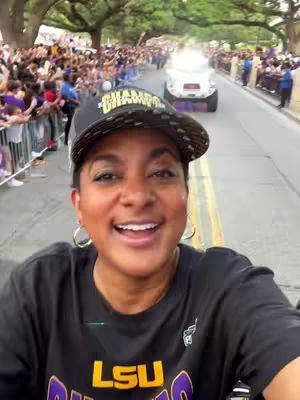 A woman smiles at the camera, positioned in front of a crowd of people and a parade float. She wears an LSU themed cap and shirt.