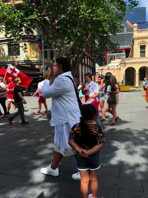 A street scene with people walking. A man in the foreground is speaking, wearing a white shirt and a black strap. A child is standing, facing the camera on the right. People and flags are visible in the background.