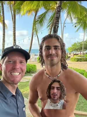 Two men are taking a selfie outdoors. The man on the left is smiling and wearing a black baseball cap. The man on the right is shirtless, wearing a necklace, and smiling. A smaller image of the shirtless man is placed over the man on the right.