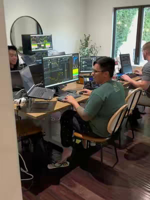 A well-lit room with multiple people working at desks with computer monitors displaying financial charts. One man is in the foreground, wearing a green shirt and glasses, actively using a computer. The setup suggests a day trading or financial analysis environment.