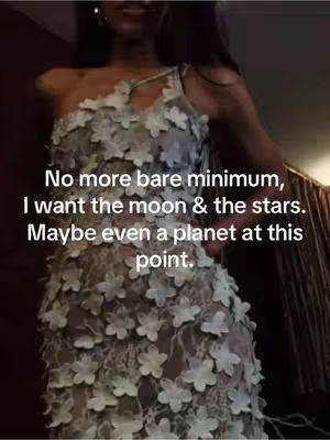 A woman in a floral-appliquéd dress stands indoors, seen from the mid-chest up. Text overlayed appears in white with a black outline against the image. Subtle lighting suggests a room setting, with darker areas, adding depth the visual scene enhances the focus on the woman and her attire.