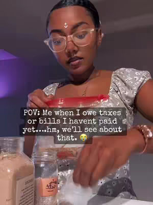 A woman with dark hair and glasses is holding a clear plastic bag, appearing to be in front of a counter or work surface. Text overlayed says, 'POV: Me when I owe taxes or bills I havent paid yet....hm, we'll see about that.'