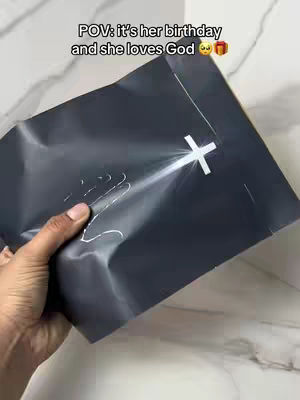A hand holds a dark, wrapped gift with a white cross design against a light-colored marble background. Text at the top reads 'POV: it's her birthday and she loves God'.