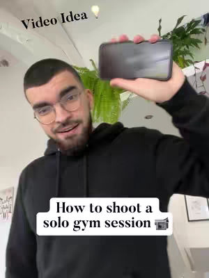 A man in a black hoodie holds a smartphone, looking at the camera and smiling. A text overlay reads "How to shoot a solo gym session". Additional text overlay reads Video Idea on the top left. The background features a light ceiling, and some plants and a painting.