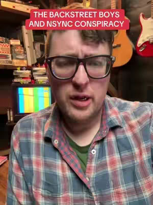 A man with glasses is speaking to the camera, with a red banner that says 'the backstreet boys and nsync conspiracy'. There is a variety of items in the background, including a guitar and some books.