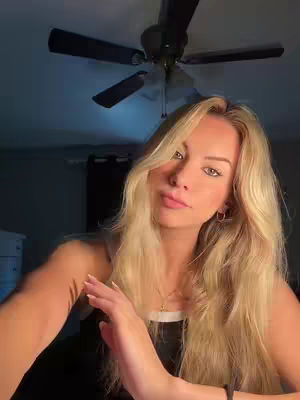 A well-lit selfie featuring a woman with long, blonde hair, looking at the camera. She is wearing a black top and a delicate necklace. The background includes a ceiling fan and a dimly lit interior, suggesting a room.