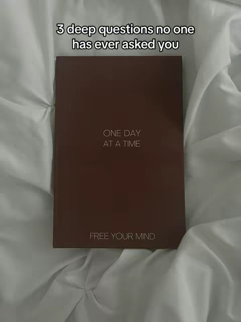 A brown journal lies open on a wrinkled white bedsheet. The journal's cover has white text, and the overall composition is simple and centered. The lighting appears soft and natural.