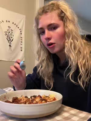 A woman with long blonde hair is holding a small, blue object and looking at the camera. She is wearing a casual black top, and a bowl with food is in front of her. A banner with text and floral design hangs in the background.