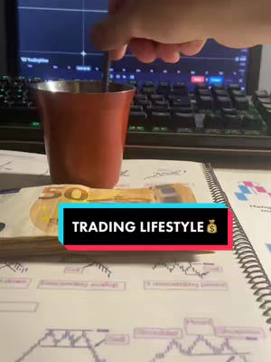 The image shows a mug, cash and a notebook with what appears to be trading charts. On the bottom there's a text label that reads 'TRADING LIFESTYLE'. Behind it is a computer screen with graphs. The composition is well-lit, slightly angled, creating a casual and inviting feel.